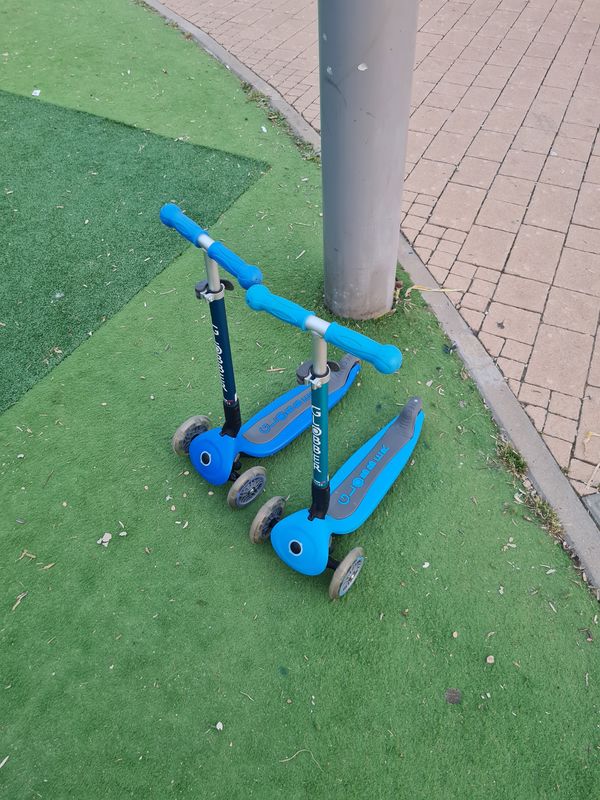 שני קורקינטי ילדים כחולים עם שלושה גלגלים עומדים על דשא סינתטי בגינת שעשועים