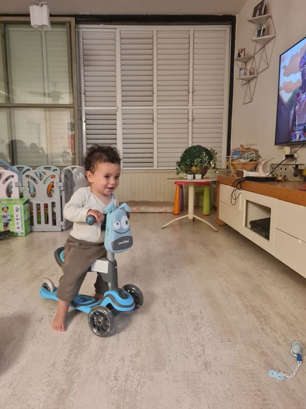 פעוט רוכב על קורקינט תלת גלגלי כחול עם מושב בסלון הבית
